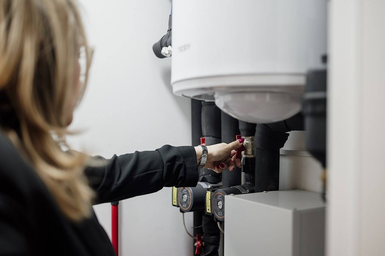 A person is adjusting a valve in a utility room where plumbing equipment is located. This action is part of routine maintenance in a home. Sunlight is present i